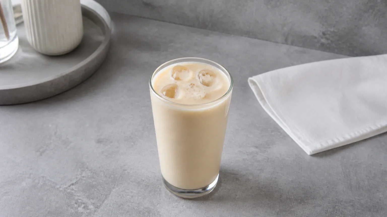 Tall glass of pale cold protein shake with ice on matte grey stone, no branding