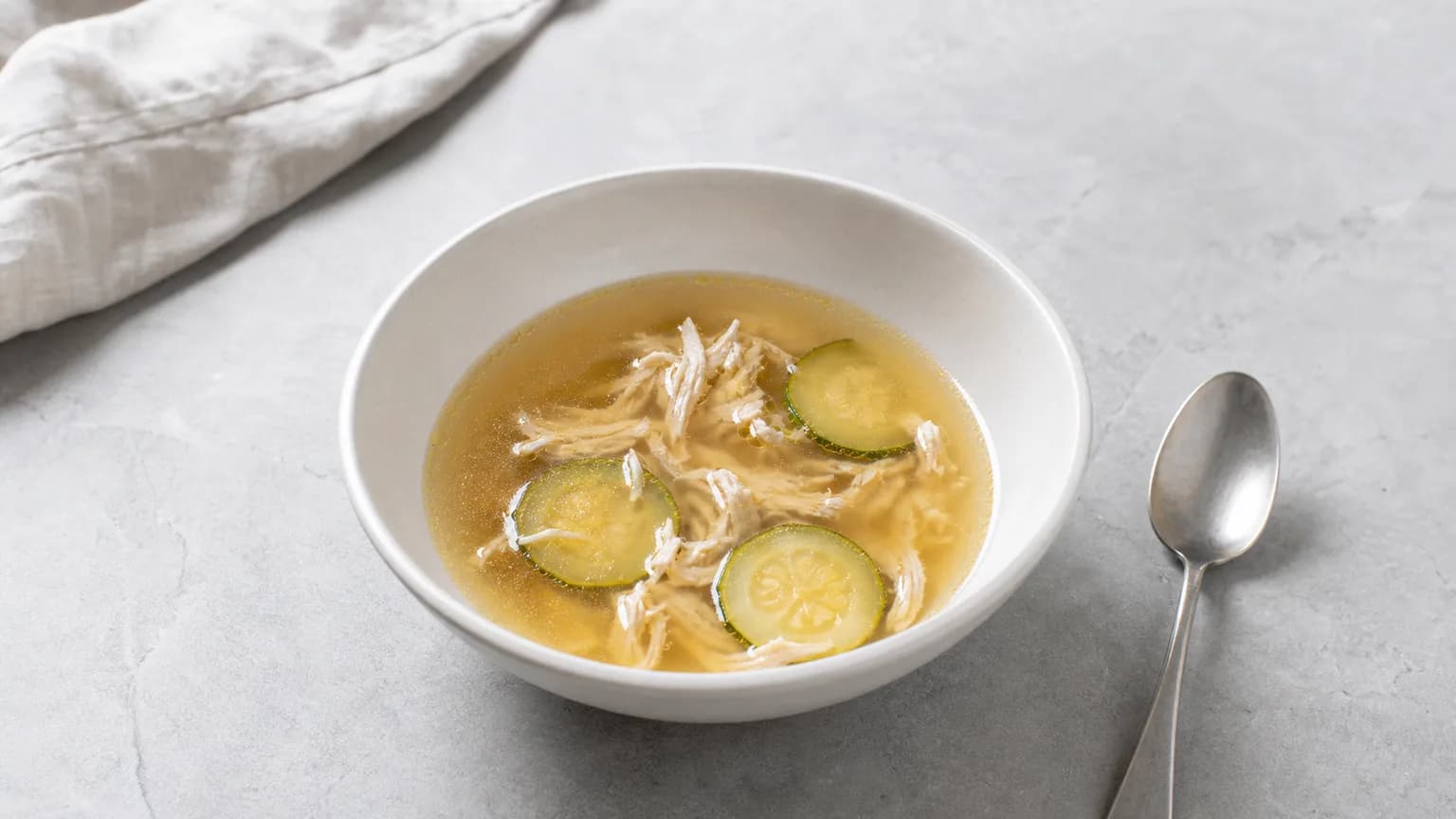 Small bowl of clear broth with a little shredded chicken and soft zucchini on grey stone