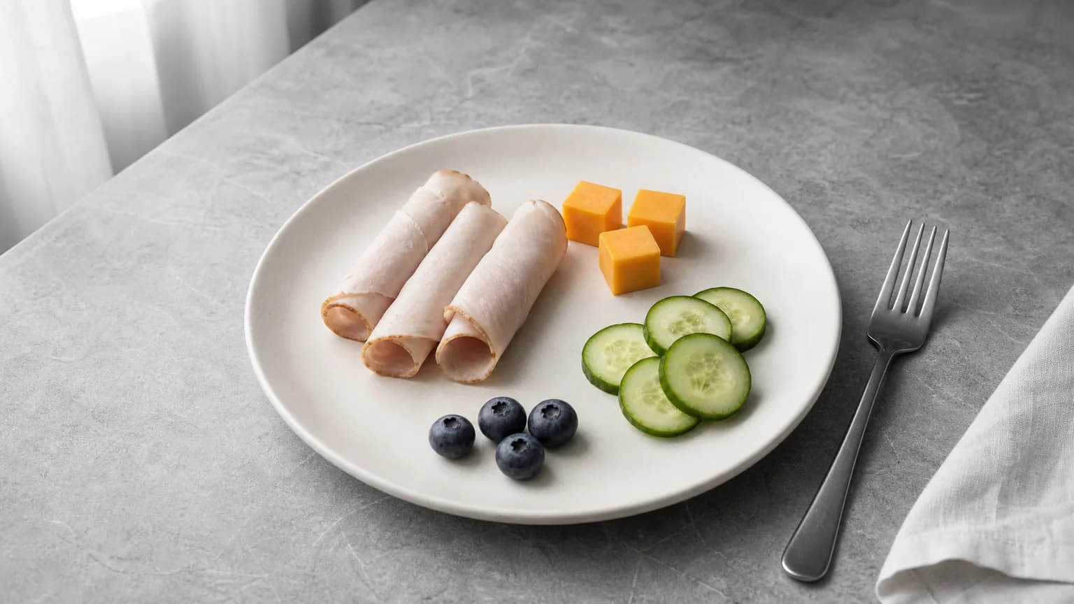Small plate with rolled turkey, cheese cubes, cucumber, and a few berries on matte grey stone
