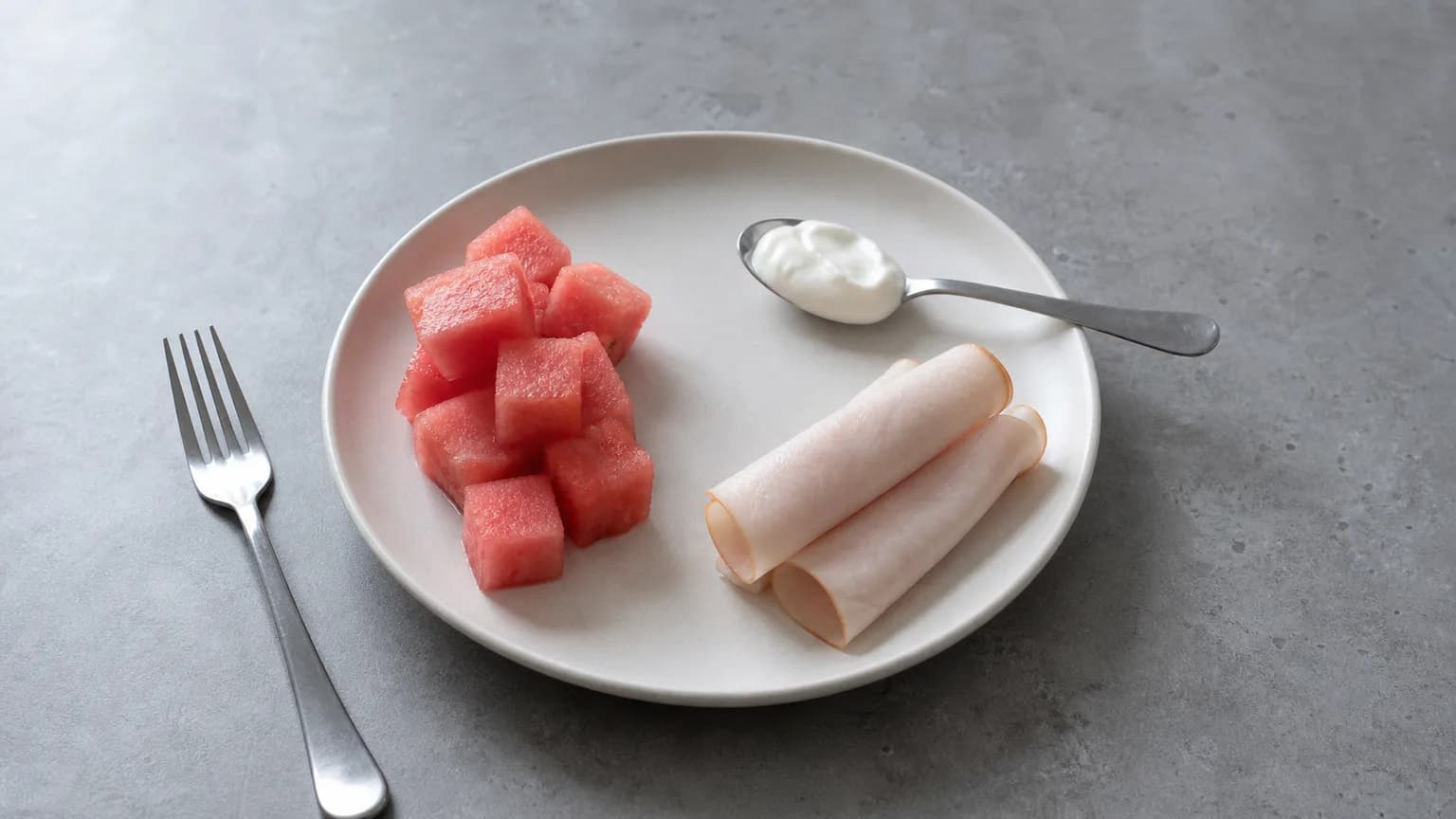 Small plate of cold watermelon cubes beside rolled turkey and a spoon of yogurt on grey stone